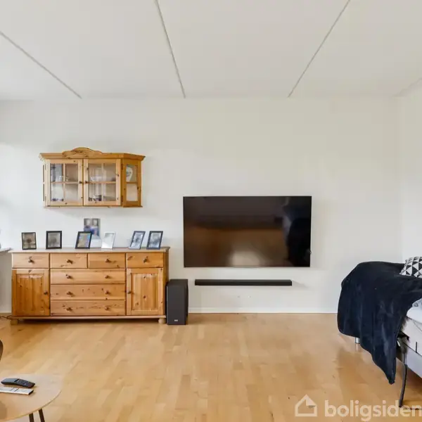 TV hænger på væggen. Under det står et skab med billeder. En seng er til højre, omgivet af sofa, radiatorskammel og lille bord i lys stue med trægulv og vindue.