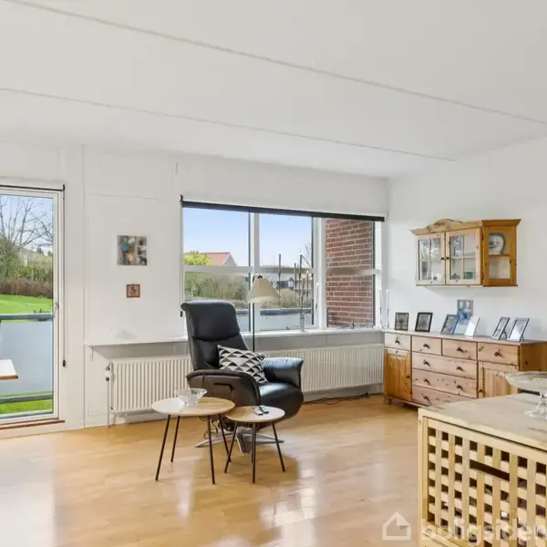 En lænestol står foran et stort vindue. Ved siden af står et lille bord. Et spisebord med stole er i nærheden, og en fladskærm hænger på væggen i en lys stue.