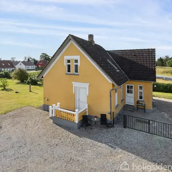 Et gult hus med sort tag står stille i en grusindkørsel. Omkring er en stor græsplæne med træer og buske. En gårdsplads er anlagt ved siden af huset.