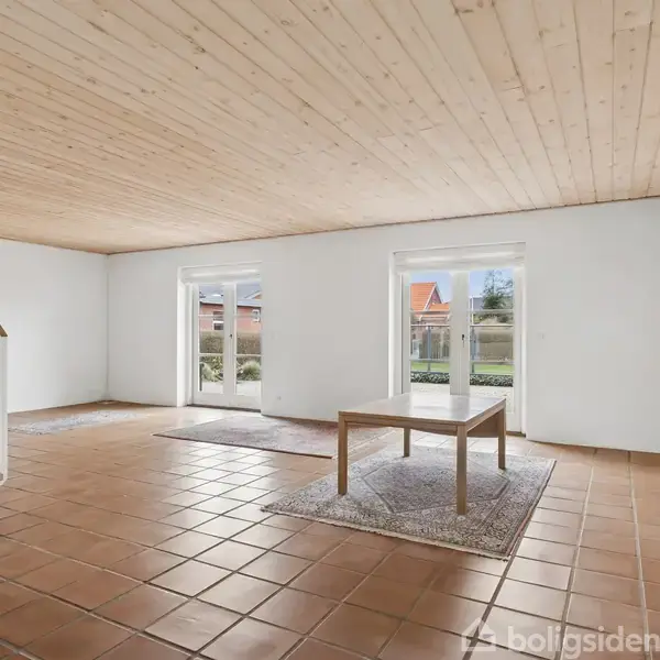 Et stort rum med et enkelt træbord på et tæppe. Lyse klinkegulve reflekterer solen, der strømmer ind gennem terrassedøre. En trappestruktur er synlig til venstre.