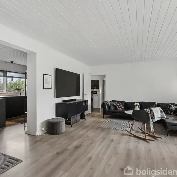 Stue med sort sofa, sofabord og fladskærms-TV på væggen. Lysindfald fra store vinduer; åbning til køkken i baggrunden. Moderne indretning med trægulv og hvidmalet loft.