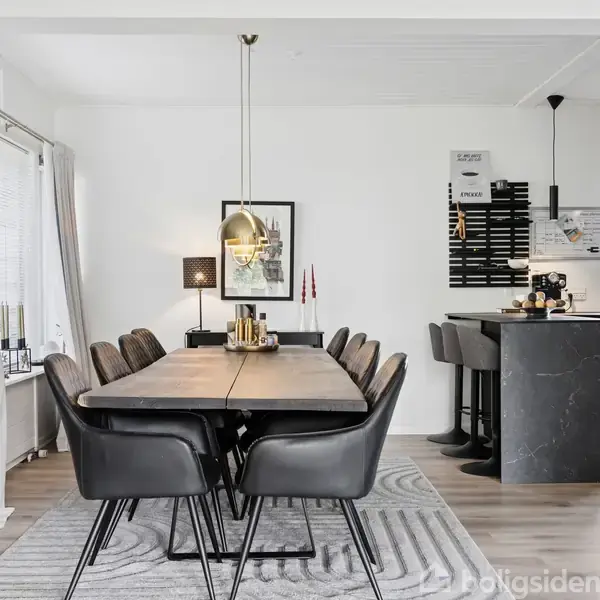 En spisestue med et langt træbord, omgivet af otte sorte stole, står centralt. En pendellampe hænger over bordet. I baggrunden ses et moderne køkken med barstole.