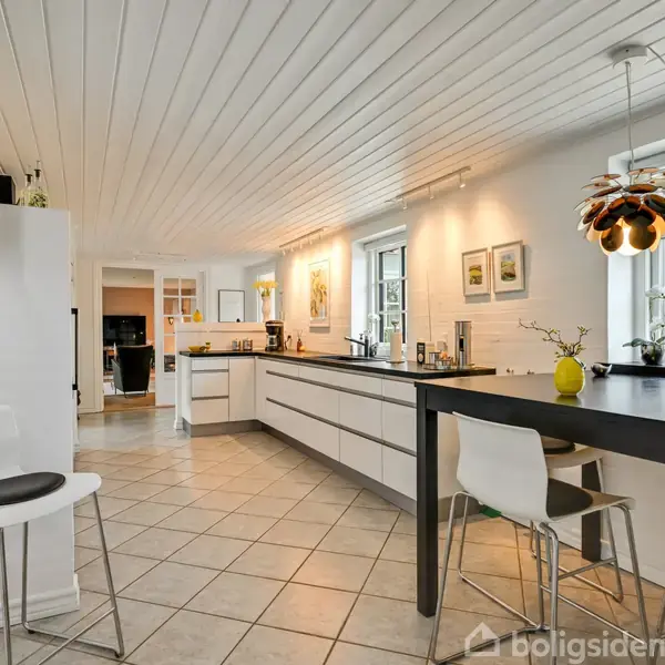 Et moderne køkken med hvide skabe og sorte bordplader, der står ved et stort vindue. Et sort spisebord med tre stole står i rummet. Lyst flisegulv og dekorative lamper.
