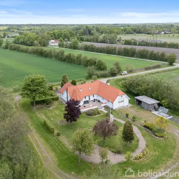 Et hvidt hus med rødt tag står omgivet af grønne marker og træer. En have med snoede stier og buske omkranser huset i et landligt landskab.