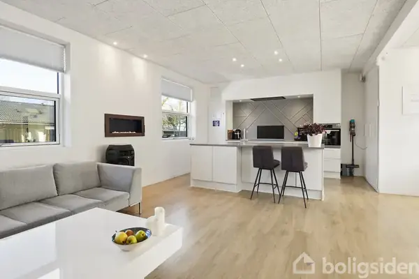 En moderne stue-køkken-alrum med en grå sofa og hvidt sofabord med frugtskål, to barstole ved køkkenøen og minimalistisk indretning, omgivet af hvidmalede vægge og trægulv.