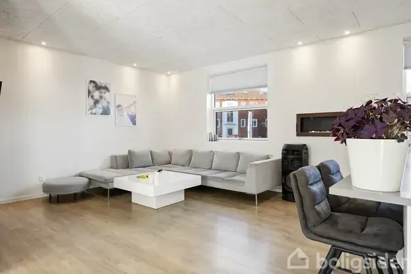 Grå hjørnesofa står i en lys stue med trægulv. Et hvidt sofabord har en gul pyntegenstand. Vægkunst hænger over sofaen. Store vinduer slipper lys ind. En plante står på bordet.