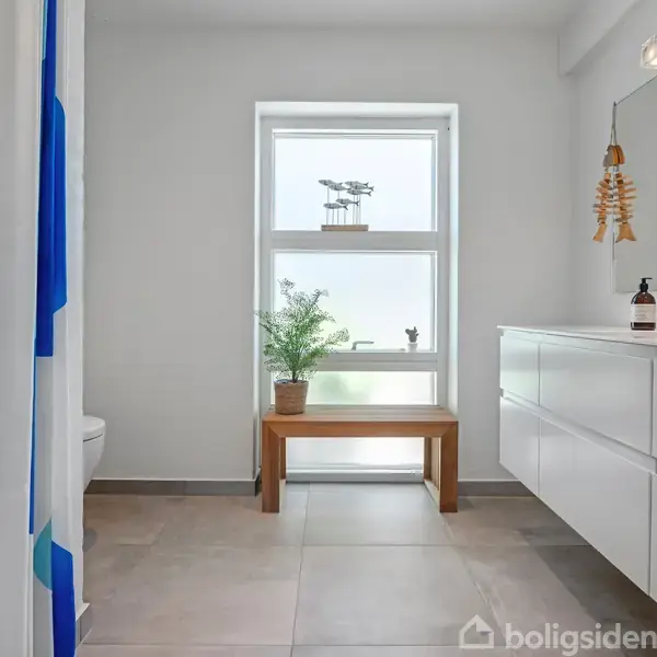 Badeværelse med hvidt bord og vask til højre. En bænk med plante foran et stort vindue. Lys indretning med fliser på gulvet.