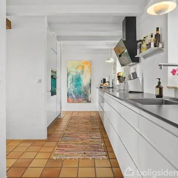 Et moderne køkken med hvidt skabsmodul langs væggen, dekoreret med planter og køkkenudstyr. Farverig abstrakt kunst hænger for enden af det flisebelagte gulv. Håndvask under vindue.