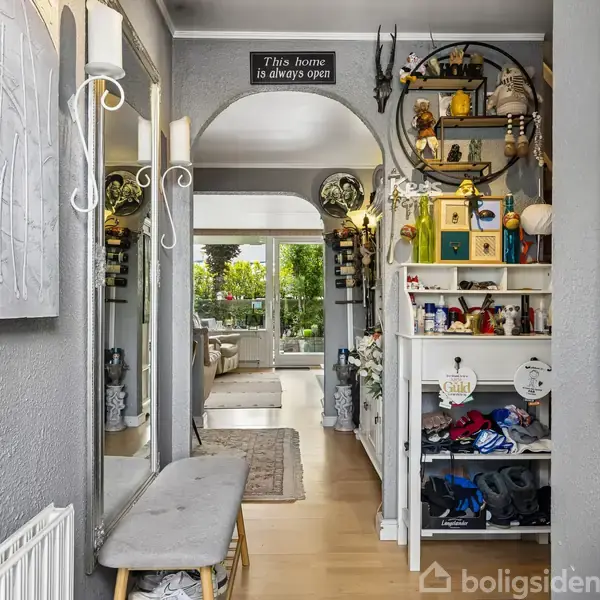 Døråbning fører til en lys stue med grønne planter. Hylder til venstre indeholder dekorationer. Ovenfor hænger skilt: "This home is always open." Skohylde til højre.