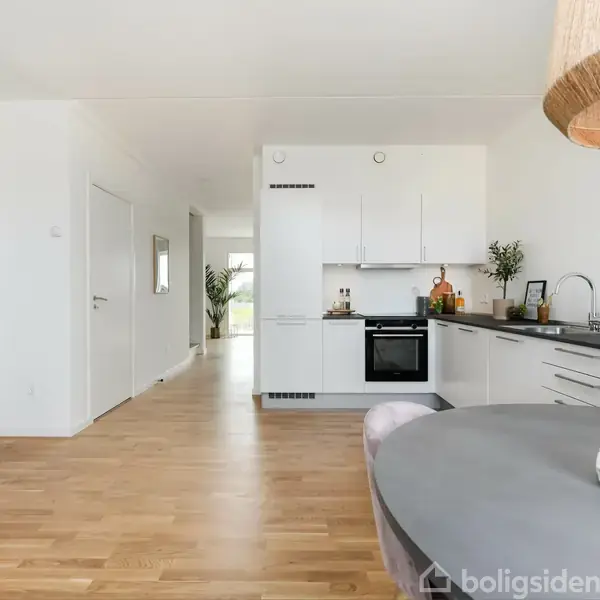 Køkkenet har hvide skabe og en sort bordplade, mens et rundt spisebord står til højre med en lysestage. Lyse trægulve fremhæver det åbne og lyse rum.