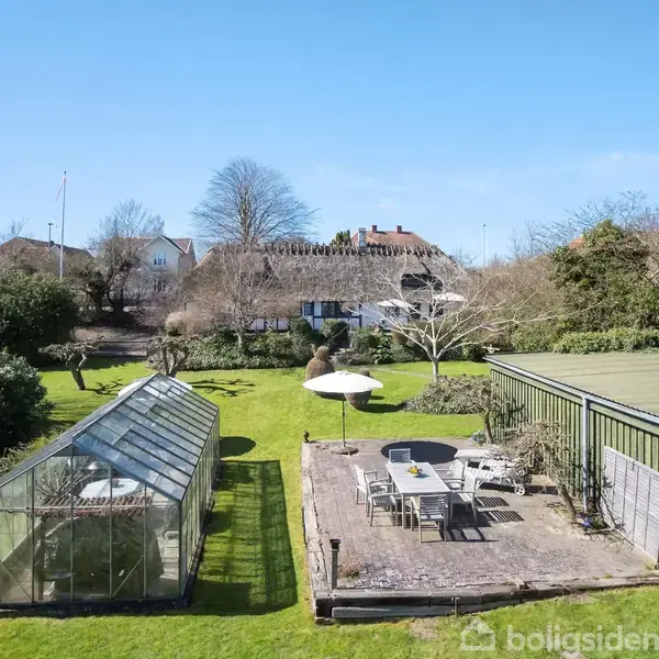 Drivhus og terrasse med havemøbler står i en velplejet have. Terrassen har en parasol, og omliggende træer markerer ejendommens grænser. Husene i baggrunden er omgivet af solen.