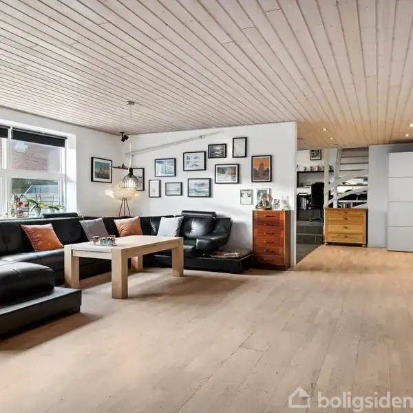 Sort lædersofa står i en moderne stue med træloft og -gulv, omgivet af vægkunst og en reol. Et træbord står foran sofaen. Rummet er lyst og rummeligt.