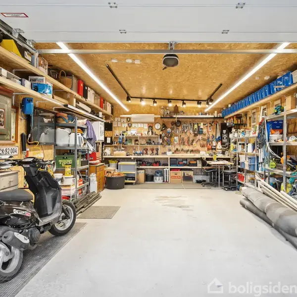 Scooter parkeret i organiseret garage; hylder med værktøj, mapper og diverse genstande langs væggene; to arbejdsstationer ved bagvæggen. Nummerplade: CL 52 004.