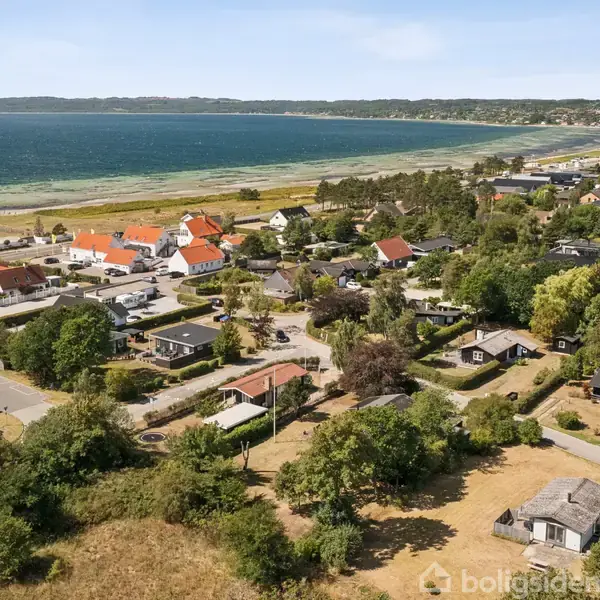 Kystlandsby set fra luften, med spredte huse og træer langs en vej. Havet strækker sig langs horisonten i baggrunden. Huse har røde tegltage, typisk for dansk arkitektur.