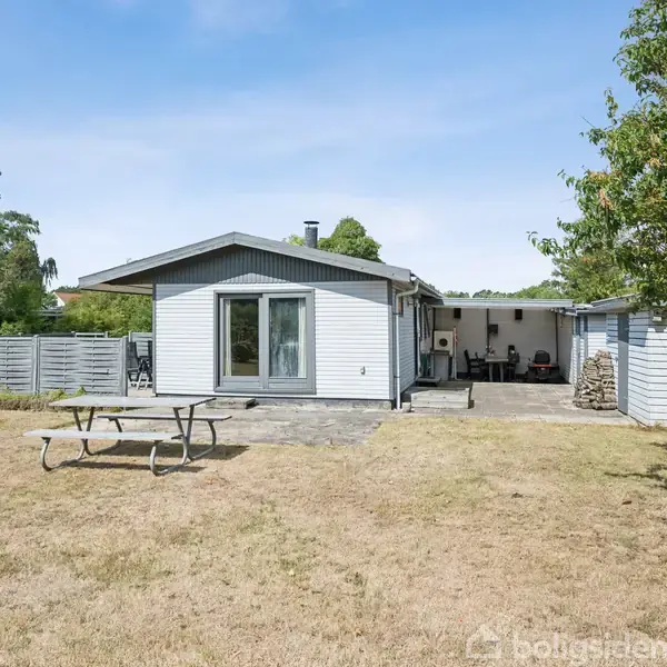Et sommerhus med træfacade står stille ved en have, hvor et picnicsæt er placeret. Omkring huset ses græs og træer, hvilket skaber en rolig atmosfære.