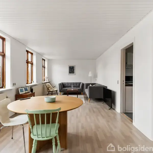 Spisebord står centralt med stole omkring, og i baggrunden ses en sofa under en lampe. Store vinduer lyser det minimalistisk indrettede rum med trælaminatgulv op. Ingen tekst er synlig.