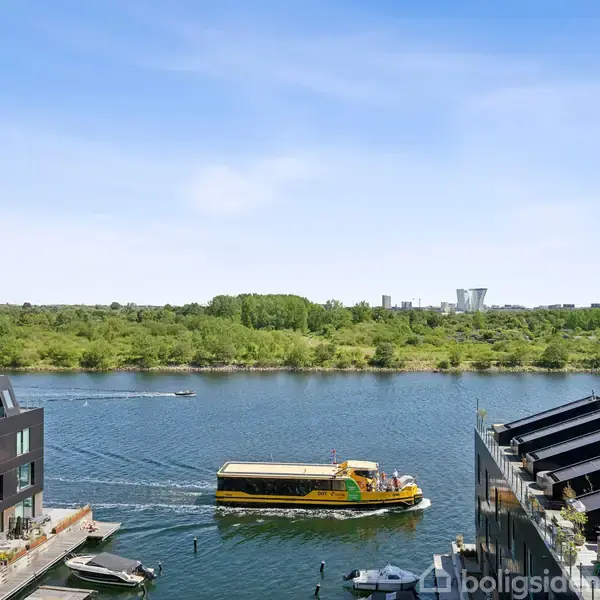 En gul båd sejler på en bred flod omgivet af moderne bygninger og grønne skove i baggrunden under en klar blå himmel.