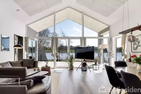 Stue med moderne møbler vender mod store vinduer, der afslører en udsigt over en sø og træer. En fladskærms-tv står foran vinduerne, mens et spisebord er til højre.