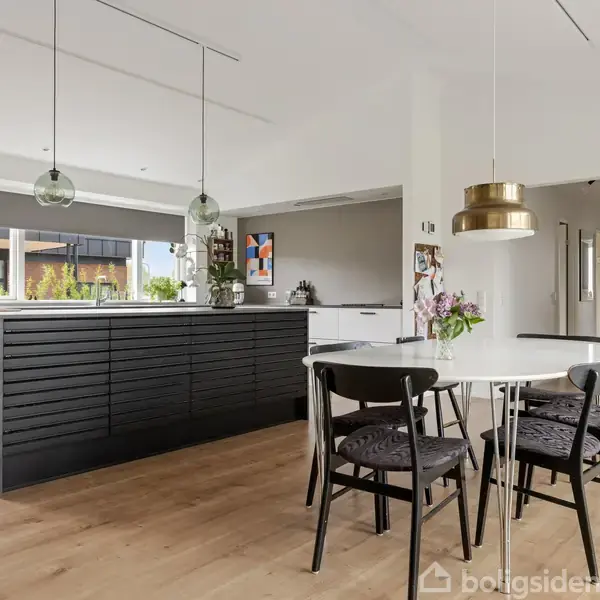 Et moderne køkken med sort køkkenø og hvide vægge. To hængelamper lyser rummet op. Rundt spisebord med fire stole. Kort på væggen og planter giver farverig atmosfære.