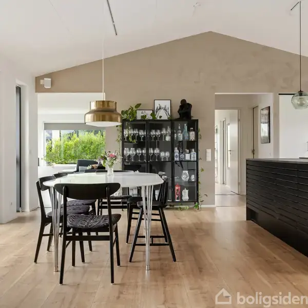 Spisebord med sorte stole står i et moderne køkken-alrum. Til venstre store vinduer, til højre en sort køkkenø. En reol med glas står foran en beige væg.
