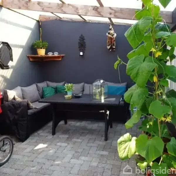 Sofa står i hjørne af overdækket terrasse, med puder og planter. Bord foran sofaen, omgivet af vægdekorationer. Rød barnevogn står til venstre, plante hænger til højre.