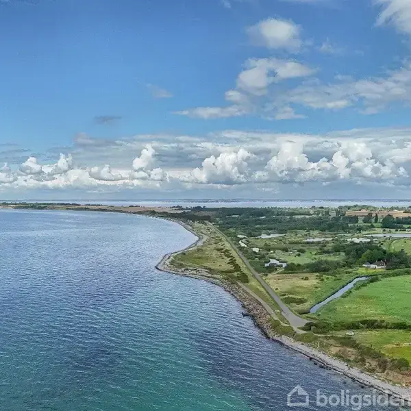 Kystlinje strækker sig langs et åbent hav, mens grønne marker og enkelte huse pryder det landlige landskab bagved, under en blå himmel med hvide skyer.