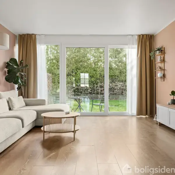 Stue med lys sofa og rundt sofabord ved store vinduer, der fører til en have med grønne buske. Et tv står på et hvidt skab til højre, omgivet af lyserøde vægge.