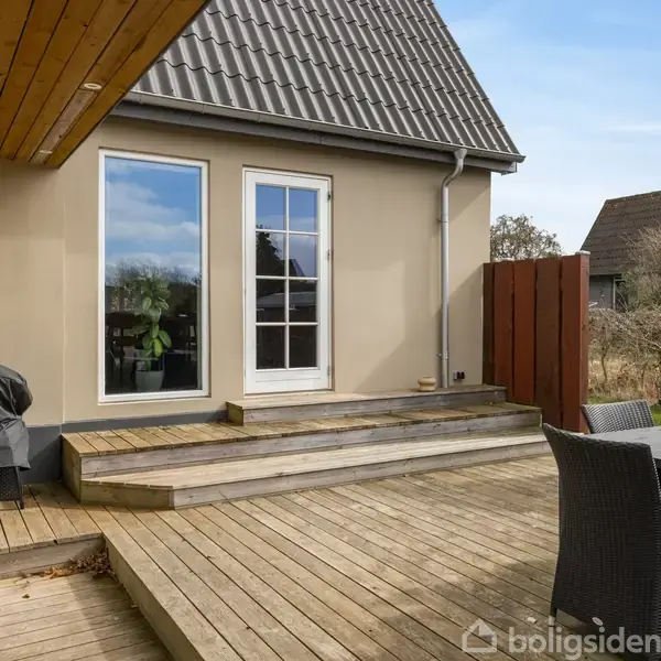Terrasse med trædæk foran et moderne hus med et skråt tag. En gasgrill står ved siden af trappen, der fører op til en glasdør. Ved siden af står et bord med stole.