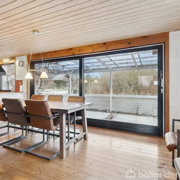 Et spisebord med seks brune stole står på et trægulv. Glasdøre åbner mod en overdækket terrasse; køkkenet ses i baggrunden med hvidt interiør og sort emhætte.