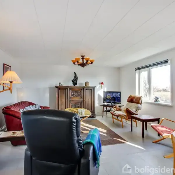 Stue med lænestole og sofaer arrangeret omkring et sofabord. En vintage skænk står ved væggen, mens et fladskærms-tv er placeret ved et vindue, der giver naturligt lys.