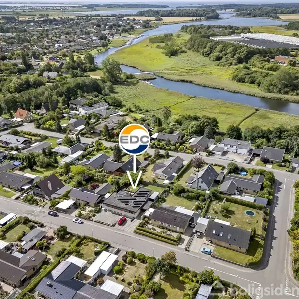 Luftfoto viser et boligkvarter med huse langs veje. En pil peger mod et specifikt hus med "EDC" logoet over det. I baggrunden ses grønne områder og en flod.