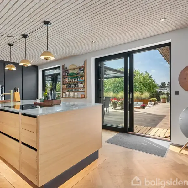 Køkkenø med sort bordplade og træskuffer står i et moderne køkken. Gulv-til-loft skydedøre åbner til en terrasse med udsigt til grønne haver.