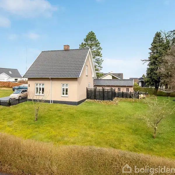 Et lille, beige hus med skråt tag står stille på en velplejet græsplæne omgivet af en lav hæk. Flere træer ses i baggrunden under en skyfri himmel.