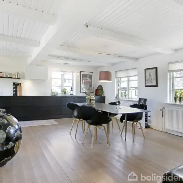 En stue med et sort spisebord og stole, omgivet af hvide vægge og træloft. Store vinduer lukker lys ind, og en stor dekoreret vase står til venstre.