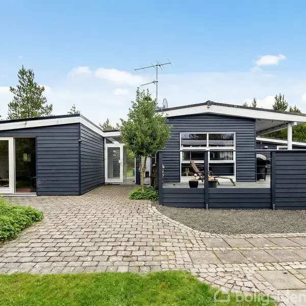 Et moderne, mørkeblåt hus med fladt tag; der er store vinduer. Omkring huset er der flisebelagt terrasse og grønne træer i baggrunden under en blå himmel.