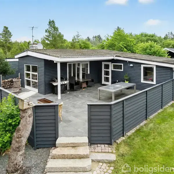 Et sommerhus med sort træfacade står på en flisebelagt terrasse, omgivet af en lav sort træhegn. Træer og buske omkranser området, skaber en frodig, privat atmosfære.