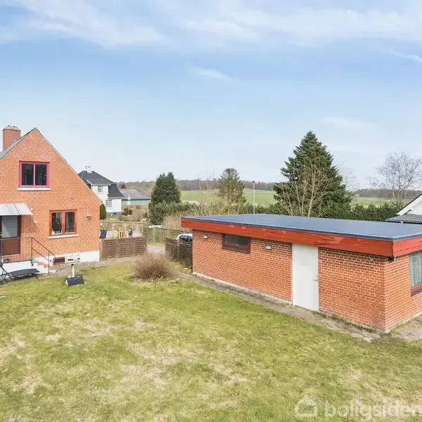 Et rødt murstenshus med trappe står på en grøn græsplæne. Ved siden af er der en rød murstensgarage. Omgivelserne inkluderer flere træer og et nabohus i baggrunden.