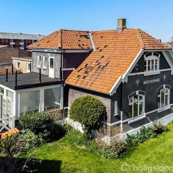 Hus i villa-stil med rød teglstenstag, store vinduer og en veranda. Teksten "Hedvigsminne" ses på facaden. Omgivet af en grøn have med buske og tættere bebyggelse i baggrunden.