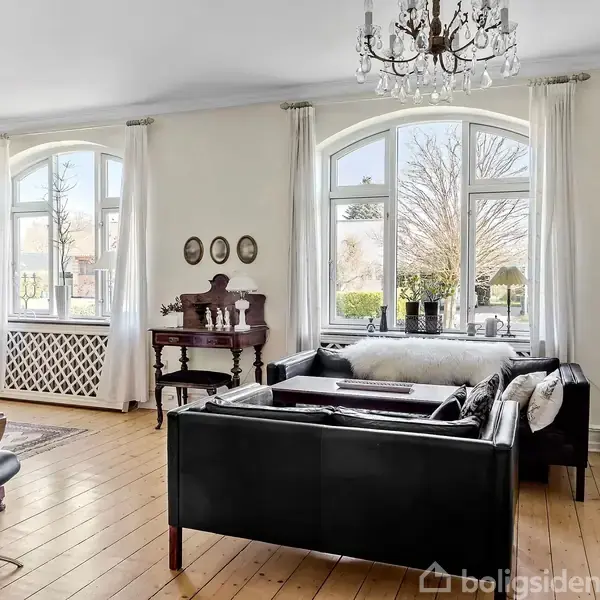 En stue med sorte sofaer står i et lyst rum med trægulve og store vinduer med udsigt. En lysekrone hænger fra loftet, og hylder fylder højre væg.