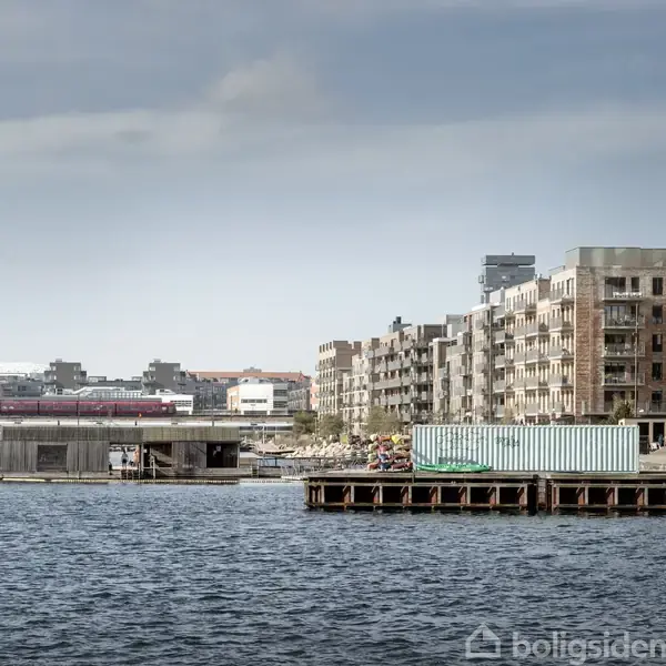 Vandfront med boliger i baggrunden. Bygningerne strækker sig langs kysten, mens en blå container og en træbro befinder sig i forgrunden. Himlen er overskyet.