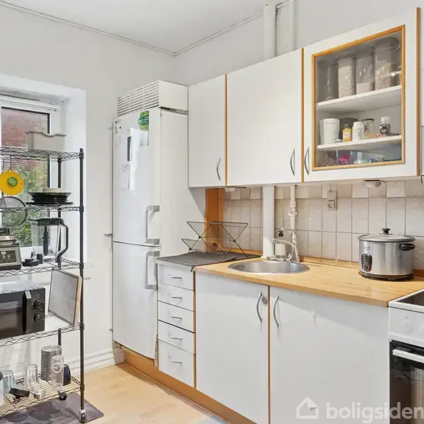 Et køkken med hvide skabe og en træbordplade, udstyret med en mikrobølgeovn, komfur, og opbevaringshylde. Køleskabet står ved siden af en væghylde med krukker. Gulvet er lyst trægulv.