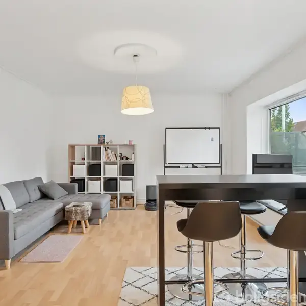 Grå sofa står stille ved siden af en reol fyldt med bøger og genstande i en lys stue med trægulv; et højt sort køkkenbord med tre stole i forgrunden.