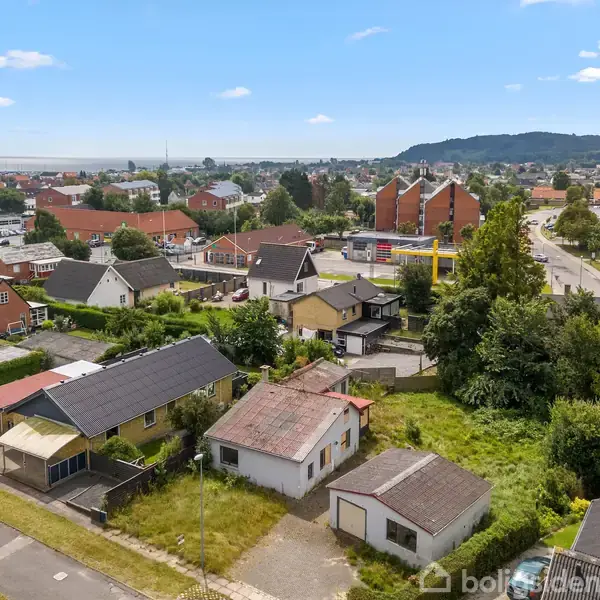Boliger tæt sammen med farverige tage i en forstad som kigger mod en vej og grønne områder, omkranset af bakker i baggrunden under en næsten skyfri himmel.