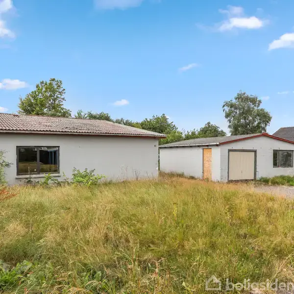 Et gråt hus med tilhørende garage står på en stor, uplejet græsplæne. Foran huset er et skilt med teksten: "Til salg." Cloudy himmel og træer i baggrunden.