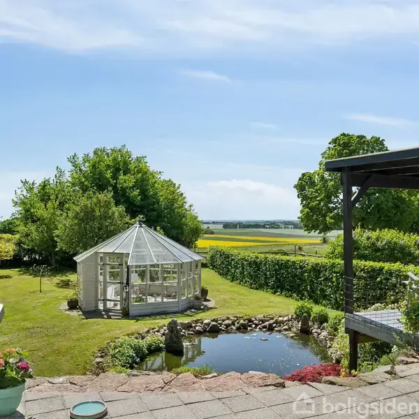 Et drivhus står stille i en velplejet have med grønne buske og træer omkring. I forgrunden ses en lille dam og en terrasse med udsigt over det åbne landskab.