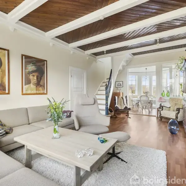 En beige hjørnesofa står i en stue med trægulv og træloft, flankeret af moderne kunstværker på væggen. Et hvidt tæppe dækker gulvet nær et sofabord.