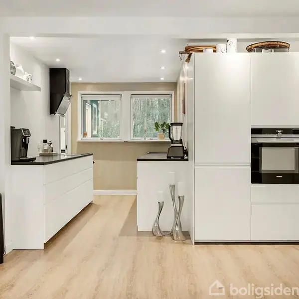 Køkken med hvide skabe og indbygget ovn. Bordplade med kaffemaskine til venstre. Vinduet i baggrunden viser træer. Trægulv i lys tone.