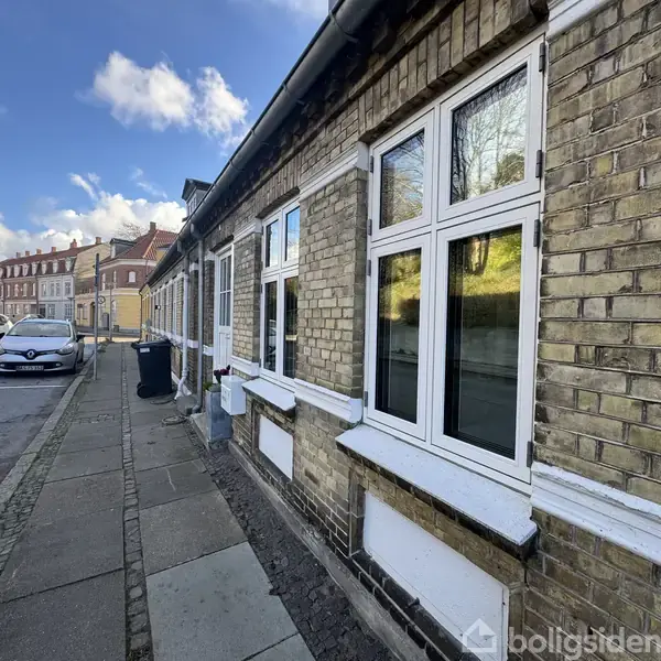 En række af gule murstensbygninger står langs en fortovskant. Vinduerne er hvide, og to biler er parkeret på gaden i solskin.