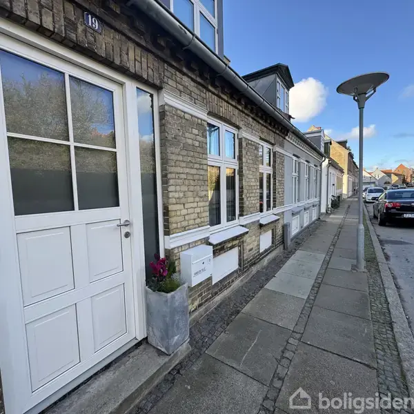Husfacade med hvide døre og vinduer står stille langs en fortovskant i en boligkvarter, hvor en gade lygte og parkerede biler er synlige i baggrunden under en blå himmel.