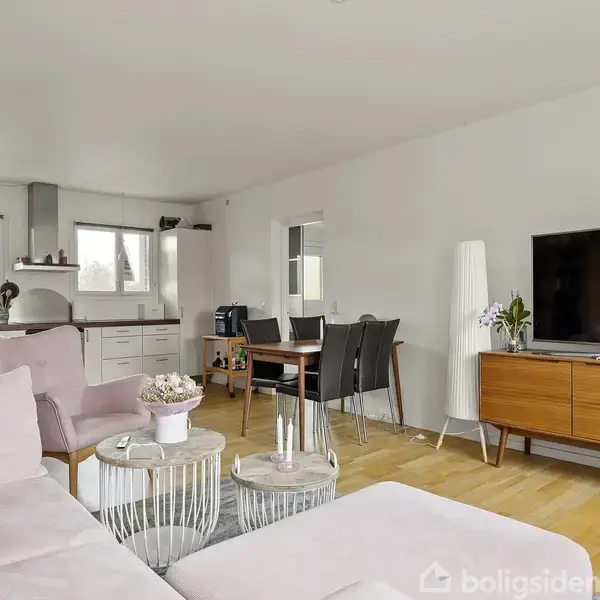 En stue har en lyserød sofa og sofabord, et højskab med TV og dekorationer, spisestuemøbler og et åbent køkken i baggrunden. Rummet er lyst med trægulv.